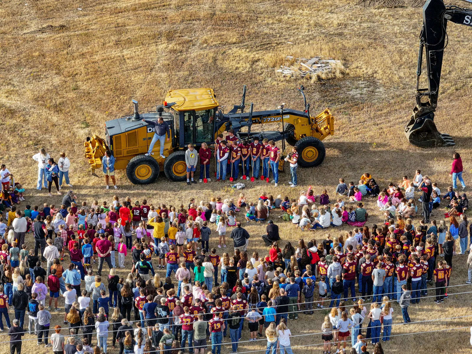 Students with red shovels stand in front of a grader as the community gathers for the groundbreaking.
