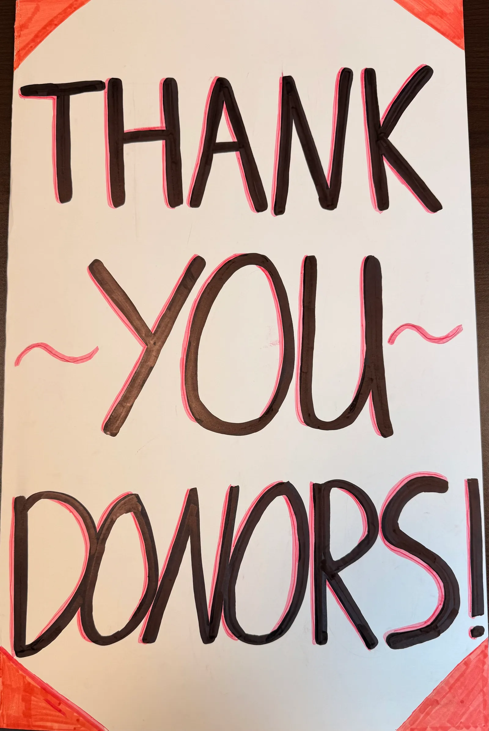A handmade sign reading Thank You Donors in black lettering outlined in pink, with red corners.